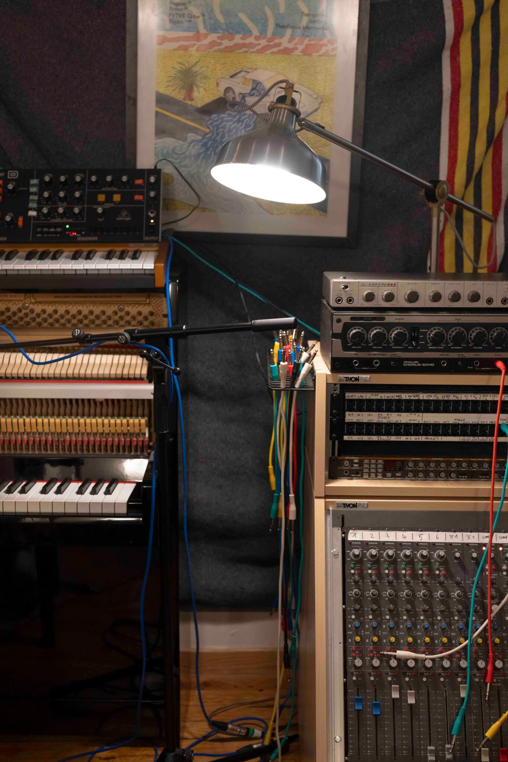 Rack de matériel analogique dans le studio de Voyou — synthétiseurs, table de mixage, chorus-echo et clavier électronique, sous une lampe d'architecte et une affiche colorée